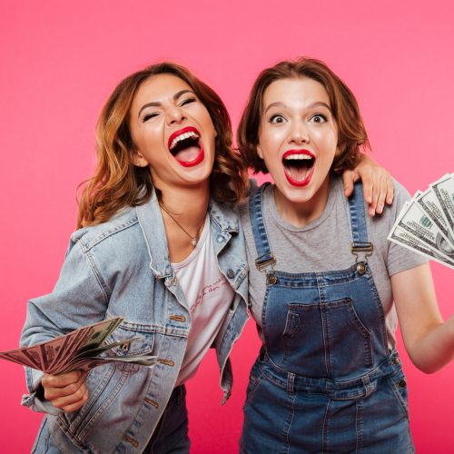 Picture of emotional pretty two women friends holding money isolated over pink background. Looking camera.